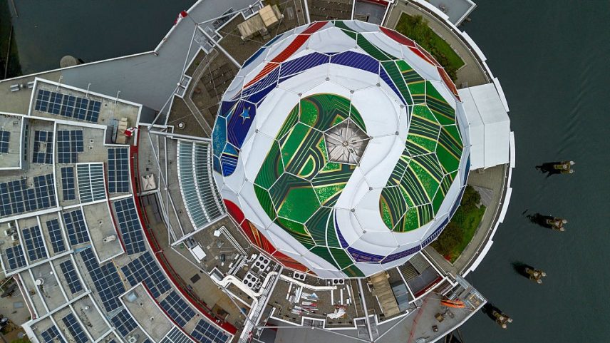Vista aérea de um edifício circular com um domo central em forma de bola de futebol, decorado com painéis verdes, azuis, brancos e vermelhos. O telhado ao redor possui vários painéis solares e há um corpo d'água escuro com barcos à direita