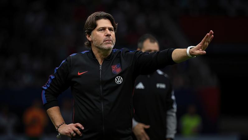 Homem de cabelo castanho e barba, vestindo agasalho preto com detalhes vermelhos e azuis, gesticula com a mão direita estendida, palma para frente, em um campo de futebol com público desfocado ao fundo
