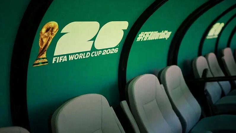 Bancos cinzas de um banco de reservas de futebol, com a parede verde-água ao fundo exibindo o troféu da Copa do Mundo e o texto "26 FIFA WORLD CUP 2026" e "#FIFAWorldCup"