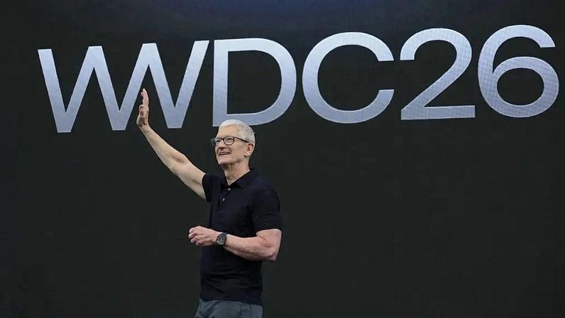 Tim Cook, CEO da Apple, sorri e acena com a mão direita levantada, vestindo polo preta e calça cinza, em um palco escuro com o letreiro WWDC26 em branco ao fundo