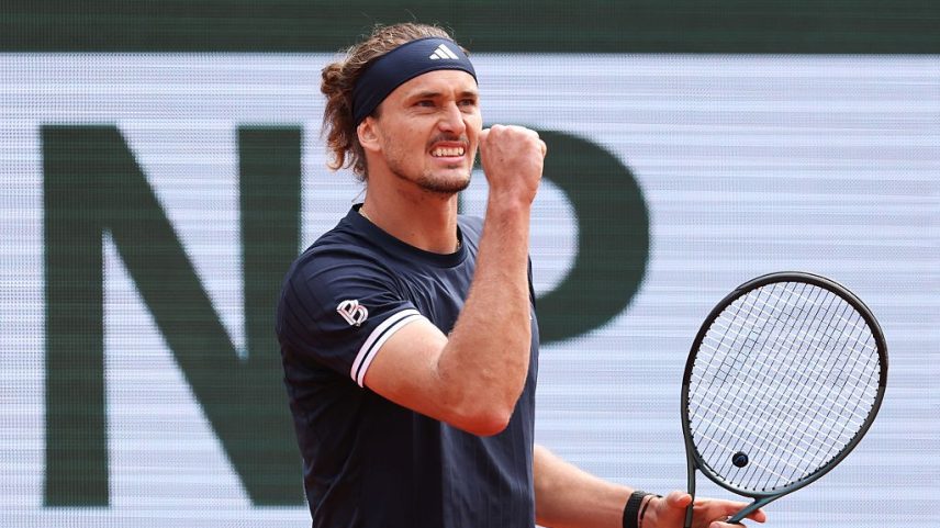 Tenista Alexander Zverev, com bandana preta e camiseta azul-marinho, celebra com punho cerrado e raquete na mão, em quadra de saibro