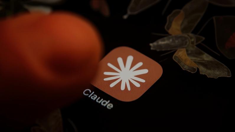 Um dedo pressiona o ícone do aplicativo Claude, que tem um fundo laranja-claro e um asterisco branco no centro, com o nome Claude abaixo