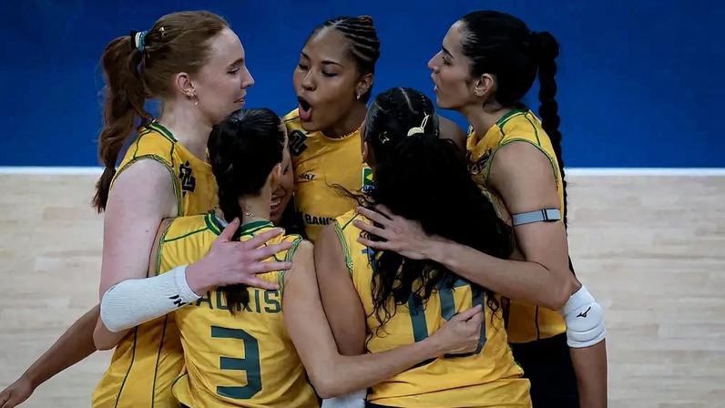 Cinco jogadoras de vôlei brasileiras, vestindo uniformes amarelos e azuis, abraçadas em quadra, com uma delas gritando de emoção