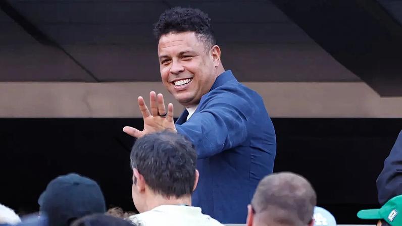 Ronaldo Fenômeno, sorridente, acena para a câmera com a mão direita aberta, usando um blazer azul e camisa branca, em meio a uma multidão com pessoas de costas