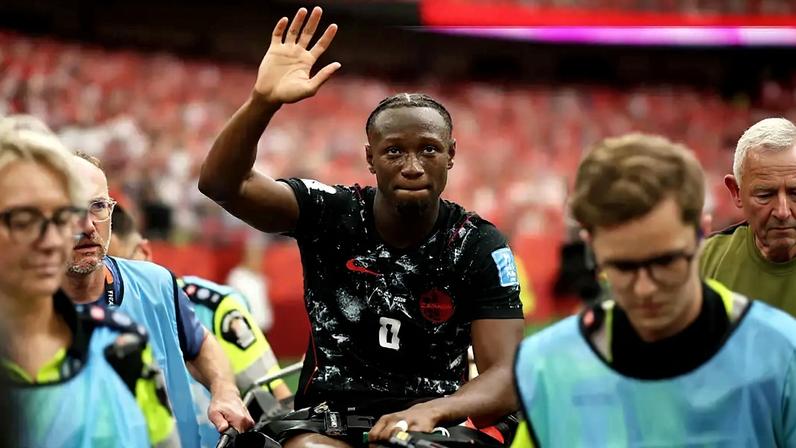 Jogador de futebol Alphonso Davies, com camisa preta e detalhes vermelhos, sentado em uma maca, acena para a torcida em um estádio lotado. Ele tem uma expressão séria, enquanto paramédicos o acompanham