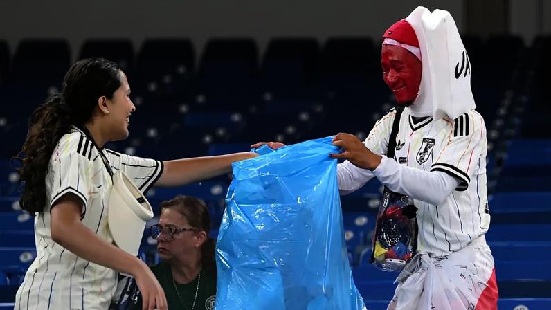 Duas pessoas em um estádio, uma mulher sorridente de camisa listrada e um homem com rosto pintado de vermelho e camisa do Japão, seguram um saco de lixo azul para recolher resíduos