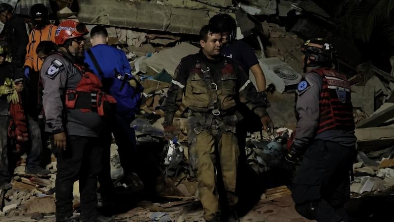 Equipe de resgate em meio a escombros de um prédio desabado. Vários homens, alguns com capacetes e coletes, caminham sobre detritos, buscando sobreviventes. Um homem de uniforme sujo de terra e colete de segurança caminha no centro, enquanto outros se movem ao redor. O cenário é de destruição, com blocos de concreto e ferros retorcidos. CARACAS, VENEZUELA - JUNE 24: Firefighters and responders search for victims in a demolished building at Los Palos Grandes after a magnitude 7.2 earthquake struck Venezuela and other regions in the Caribbean on June 24, 2026 in Caracas, Venezuela. According to the U.S. Geological Survey (USGS), the main earthquake was followed by a 7.5-magnitude aftershock less than a minute later; although some buildings collapsed, the number of victims remains unknown. (Photo by Edilzon Gamez/Getty Images)