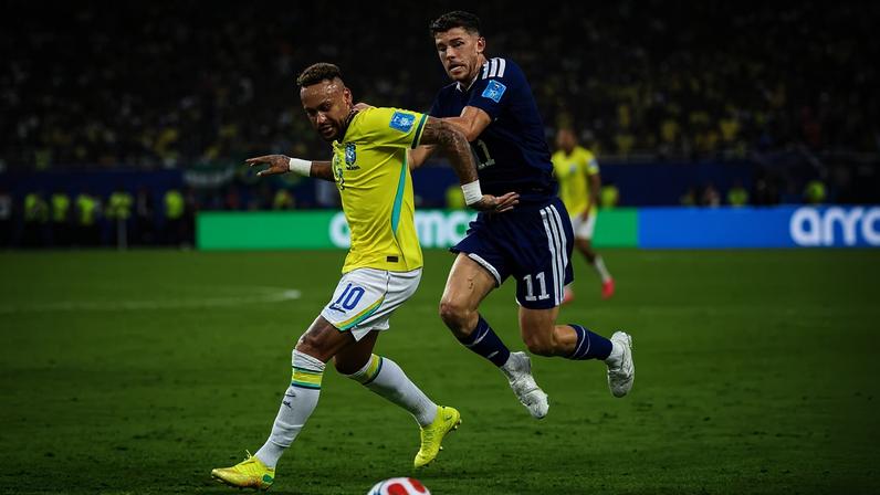 Neymar, com camisa amarela e shorts brancos, dribla um jogador de camisa azul marinho e shorts azuis, em campo de futebol com torcida ao fundo