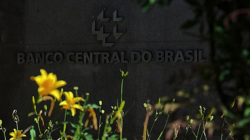 Fachada do Banco Central do Brasil com logotipo em destaque e flores amarelas em primeiro plano