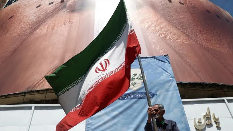 Homem segura bandeira iraniana perto de outdoor anti-EUA representando o presidente dos EUA Donald Trump e o Estreito de Ormuz em Teerã, 30 de maio de 2026.