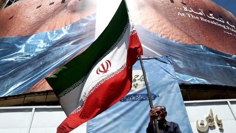 Homem segura bandeira iraniana perto de outdoor anti-EUA representando o presidente dos EUA Donald Trump e o Estreito de Ormuz em Teerã, Iraque, 30 de maio de 2026
