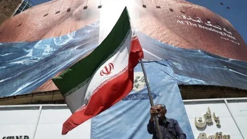 Homem segura bandeira iraniana perto de outdoor anti-EUA representando o presidente dos EUA, Donald Trump, e o Estreito de Ormuz, em Teerã, Irã, em 30 de maio de 2026