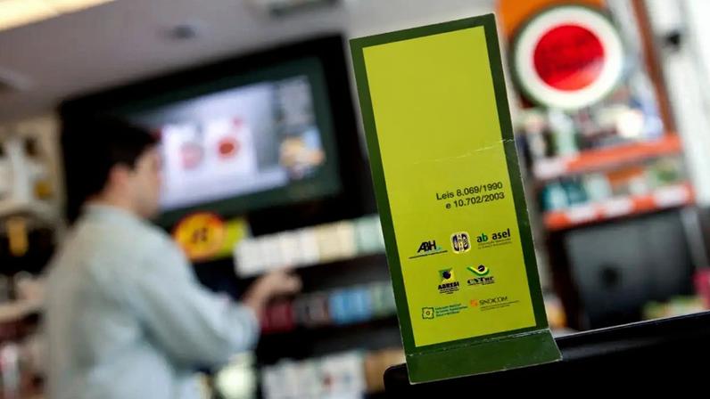 Placa verde com aviso É a lei: Não vendemos cigarros para menores de 18, com um homem desfocado ao fundo organizando produtos