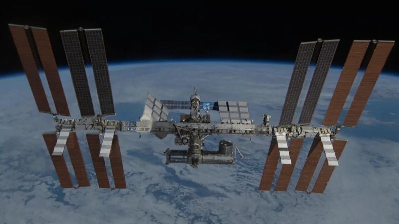 Estação Espacial Internacional (ISS) flutuando acima da Terra, com painéis solares abertos e o planeta azul parcialmente coberto por nuvens brancas, contrastando com o espaço escuro
