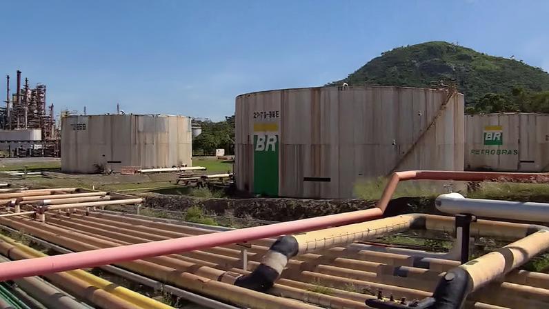 Tonéis com a logomarca da Petrobras estão no meio de um campo com diversos canos que transportam petróleo até eles.