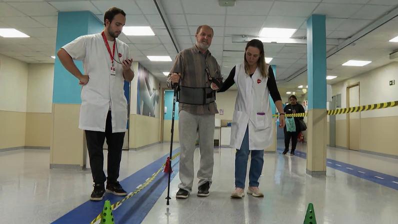 Idoso com muleta caminhando com a ajuda de dois profissionais da saúde
