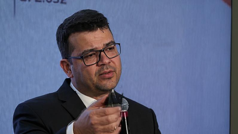 Um homem de óculos, cabelo negro e terno preto discursa com um microfone na mão e um controle de apresentação na outra.