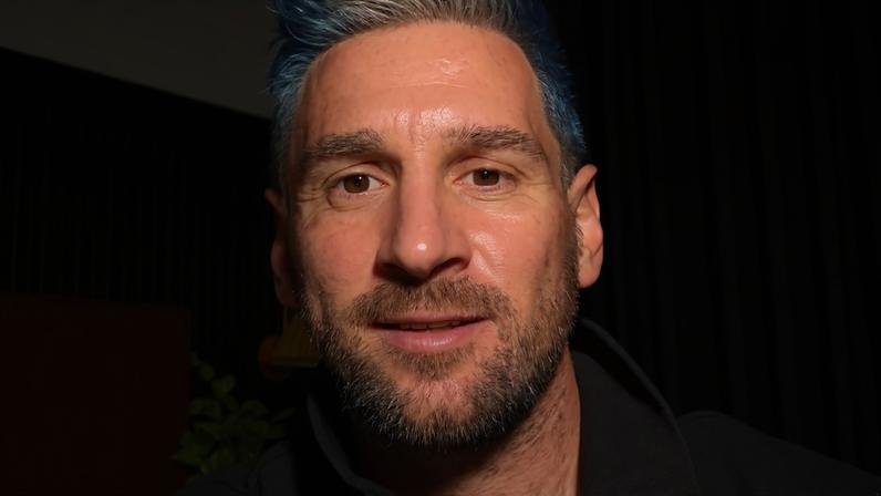 Homem com barba e cabelo azul vibrante, com uma mecha branca na frente, sorri levemente para a câmera. Veste um casaco cinza escuro com zíper, em um ambiente interno com cortinas escuras e uma planta verde ao fundo