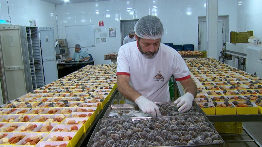 Funcionário utiliza luvas e touca ao organizar bandejas de doces artesanais em uma produção industrial, com diversas variedades alinhadas sobre a mesa.