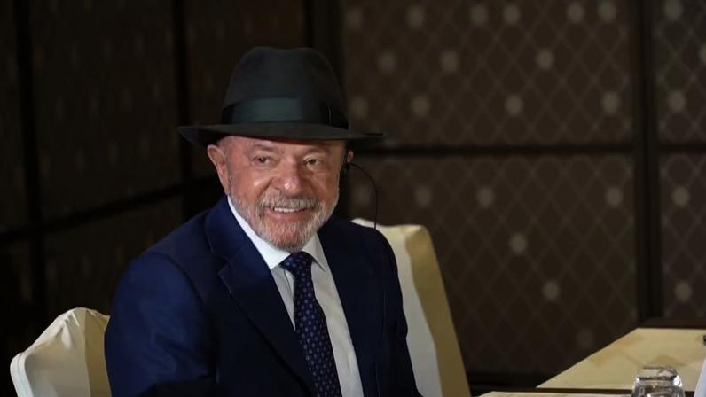 Presidente Lula, de terno azul e chapéu preto, sentado em uma mesa de reunião, com outras pessoas ao fundo