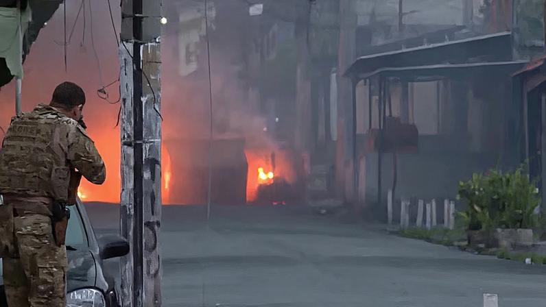 Pessoa em uniforme camuflado se protege atrás de um carro enquanto aponta uma arma. Ao fundo, fogo intenso no meio da rua gera fumaça espessa que encobre parte da cena. O ambiente urbano mostra prédios e fios elétricos cruzando o espaço.