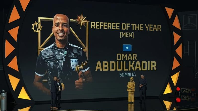 Um palco de premiação com um telão exibindo a foto de Omar Abdulkadir, árbitro da Somália, sorrindo e vestindo uniforme da FIFA. O texto na tela diz REFEREE OF THE YEAR [MEN] e OMAR ABDULKADIR SOMALIA. No palco, duas pessoas de terno entregam um troféu a um homem, enquanto outras duas pessoas, uma de terno e outra com vestimenta amarela, observam. Uma plateia assiste ao evento