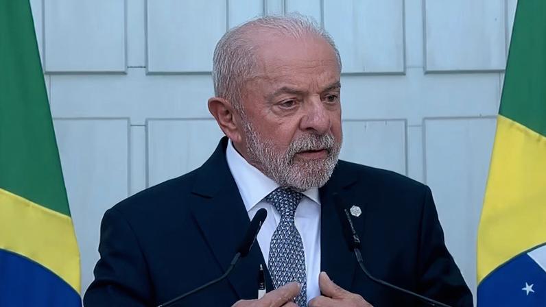 Presidente Lula, de terno, discursando em um púlpito, entre duas bandeiras do Brasil