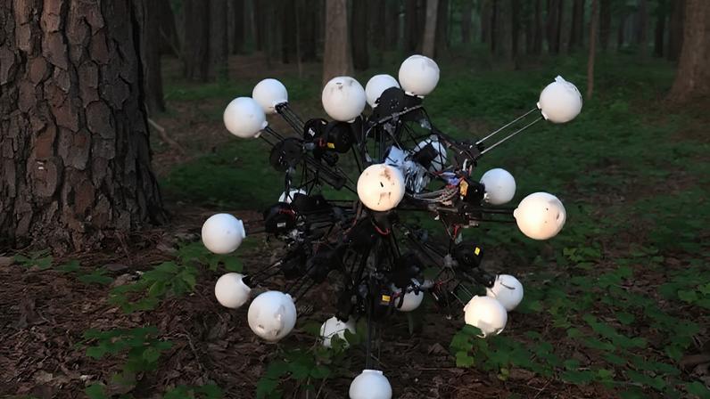 Robô esférico com estrutura complexa, posicionado em meio a uma floresta. Ele possui diversos braços mecânicos pretos, cada um com uma esfera branca na ponta.