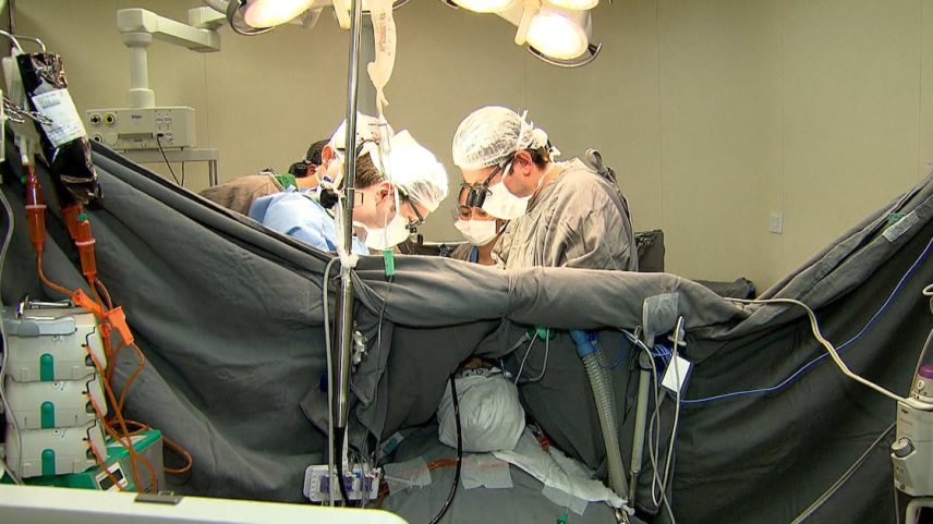 Equipe médica realizando cirurgia em sala operatória com paciente coberto e equipamentos ao redor