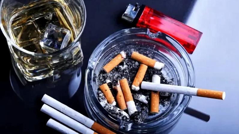 Image, vista de cima, de um cinzeiro com várias bitucas de cigarros e um copo de whisky com pedras de gelo. Ao lado vê-se um isqueiro vermelho e três cigarros intocados.