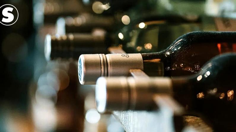 Garrafas de vinho escuras, com rótulos e tampas prateadas, alinhadas horizontalmente em uma adega escura, com pontos de luz desfocados ao fundo.
