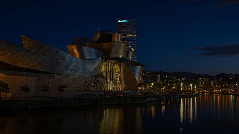 Imagem do Museu Guggenheim Bilbao, com sua arquitetura metálica curvilínea, reflete a luz do entardecer sobre o rio. Ao fundo, um arranha-céu e a cidade iluminada se estendem até as montanhas. Uma escultura de aranha gigante está em frente ao museu.