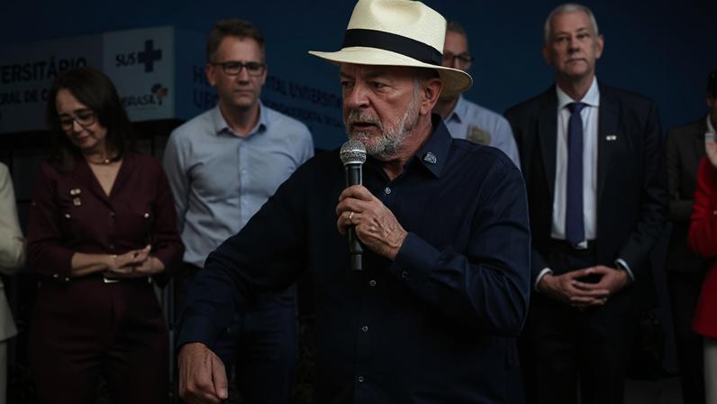 Lula de chapéu branco fala ao microfone em evento público, com outras pessoas ao fundo e painel institucional atrás