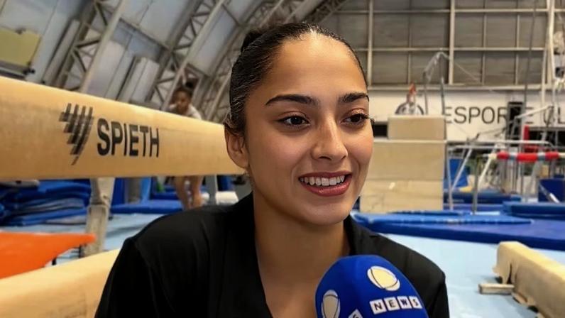 A ginasta Julia Soares sendo entrevistada dentro de um ginásio em que está acontecendo os treinos de ginástica artística. Ela está vestindo uma camiseta preta e com os cabelos presos em um coque.