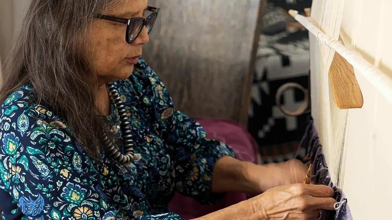 An indigenous woman with long gray hair weaves at a loom.