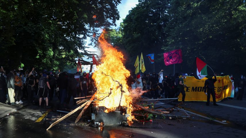 Manifestantes com bandeiras e faixas se reúnem perto de um incêndio em uma rua, em meio a confrontos com policiais antimotim durante um protesto contra a próxima cúpula do G7