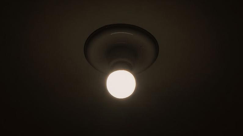 Lâmpada acesa, fixada em um soquete de teto simples, emitindo uma luz branca intensa que cria um halo circular na superfície ao redor.