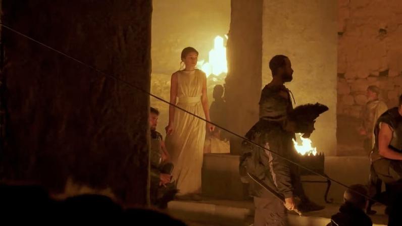 Cena de filmagem com um homem em primeiro plano e, ao fundo, uma mulher de vestido branco e cabelo preso diante de um ambiente iluminado por tons alaranjados e fogo.