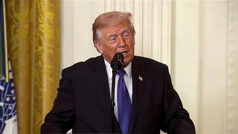 Donald Trump, de terno escuro e gravata azul, discursando em um púlpito com microfone, em ambiente interno com colunas brancas ao fundo