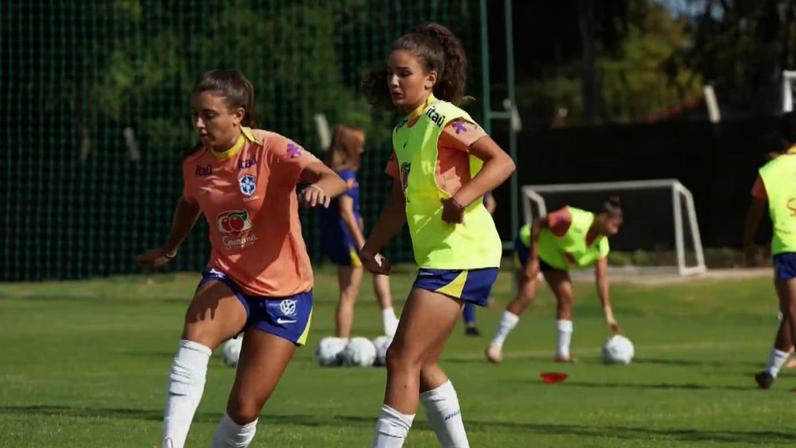 Jogadoras de futebol em treinamento em um campo gramado, sob sol e céu claro.