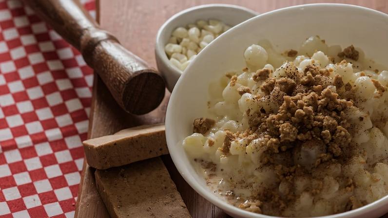 Canjica com paçoca servido em um bowl branco, decorado com canela. Ao lado, está um pote menor com grãos de canjica. Os potes estão servidos em cima de uma tábua de madeira, com duas paçocas apoiadas e um pilão de madeira. A tábua está em cima de uma toalha de mesa de estampa xadrez, de cor vermelho e branco.