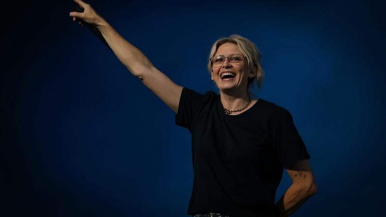 Emily Armstrong performs on stage and holds her hands up. She is wearing a black t-shirt and black belt. She has blonde hair tied up, she is wearing yellow glasses.