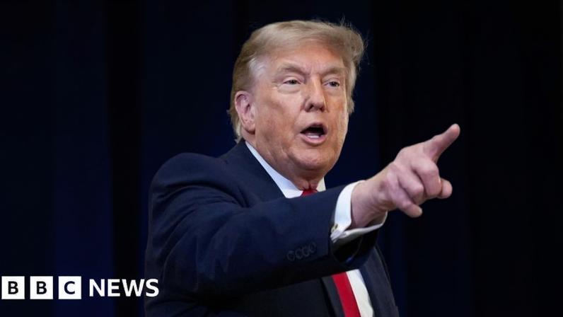 Donald Trump wearing a navy suit and red tie, points his finger to the right side of the frame.