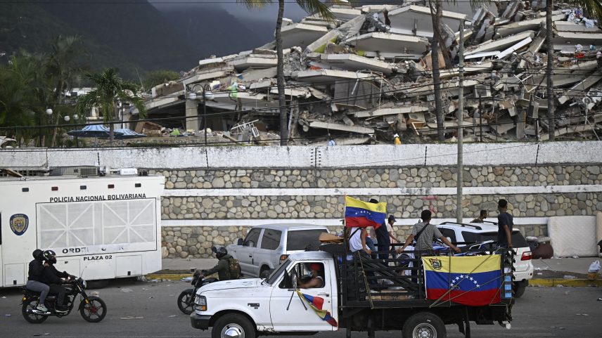 Um caminhão com bandeiras venezuelanas passa por um edifício desabado após dois terremotos, em Los Corales, no estado de La Guaira, a cerca de 40 km ao nordeste de Caracas, em 27 de junho de 2026. — Foto: FEDERICO PARRA/AFP