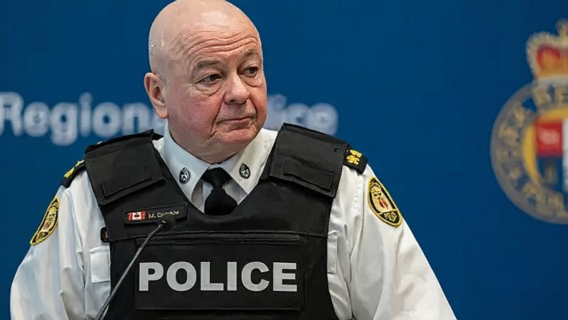 Getty Images An image of Myron Demikw, Toronto police chief, at a press conference. He is standing behind a microphone. Behind him is a blurred out blue background featuring the police emblem. He is wearing a black bullet proof vest that says POLICE on top of a white undershirt that features police patches on either arm. He is bald, and is looking at the direction of the right.