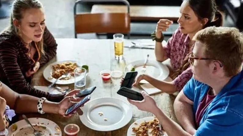 Getty Images Three young people viewed from above are sitting at a table at the end of a meal. They have their phones out and the woman on the left is frowning as she looks at hers. There are empty and half empty plates and glasses.