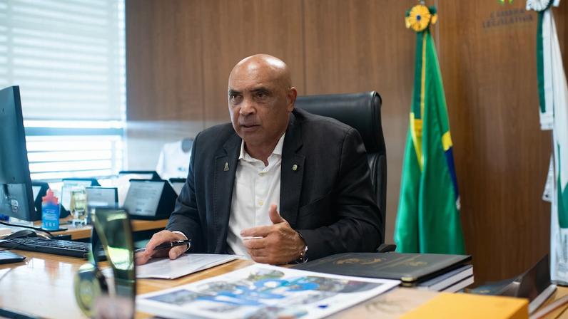 Foto de homem sentado em cadeira com mesa à frente e bandeira ao fundo