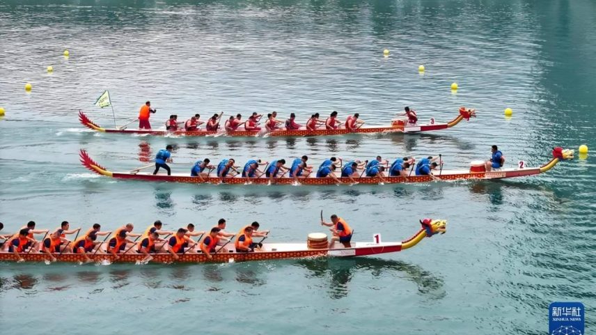 Três barcos-dragão competem em uma corrida aquática, com remadores uniformizados em vermelho, azul e laranja. Boias amarelas marcam o percurso na água verde-azulada.