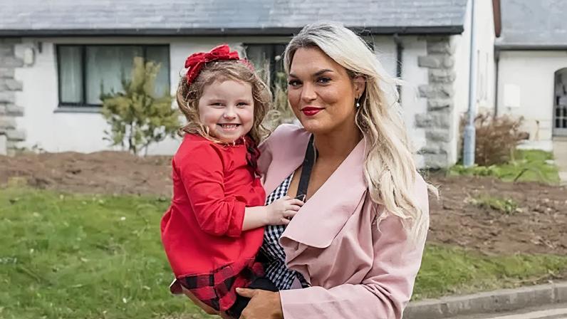 Inisowen Independent Natalie McLaughlin holds her young daughter Ella in her arms. She is smiling, has long blonde hair, and is wearing silver earrings, a pink overcoat and a black and white checked blouse. Ella is also smiling, and has a red bow in her long brown hair. The little girl is wearing a red dress. Behind them is a garden and house.