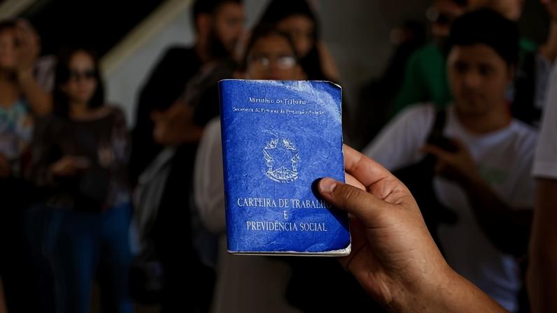 Mão segurando carteira de trabalho e pessoas passando ao fundo, desfocadas